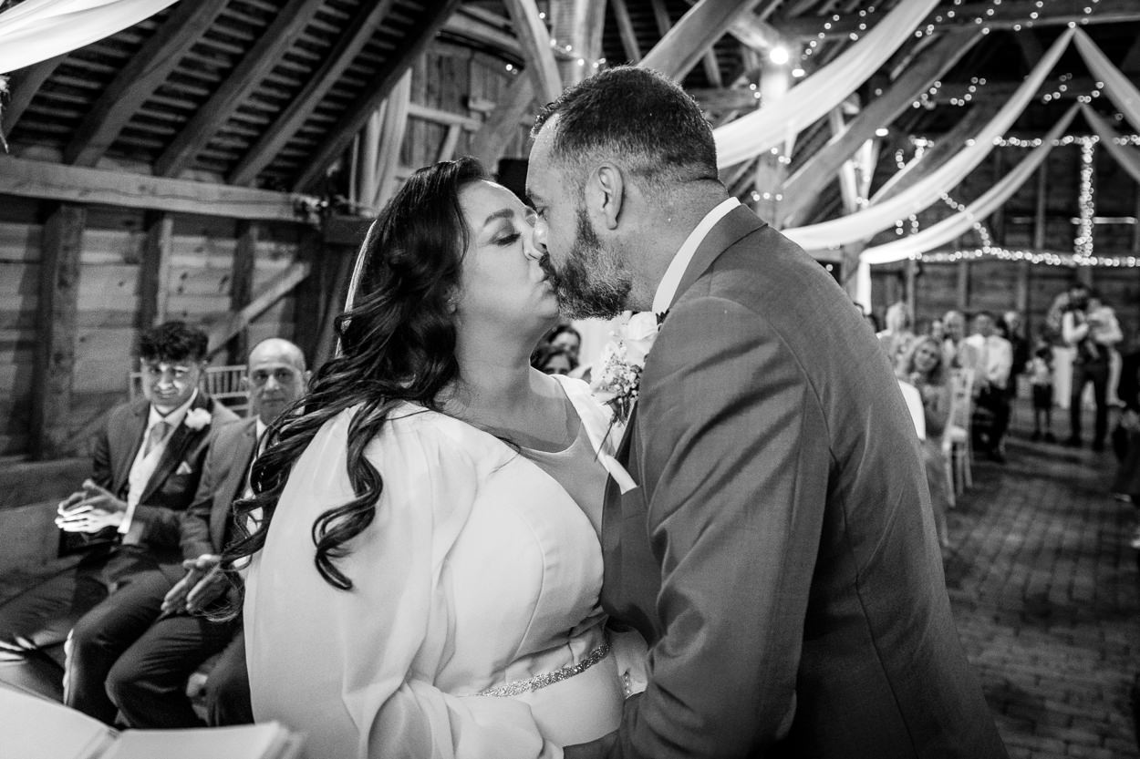 black_white image of bride and groom kissing in barn during wedding ceremony at relaxed gildings_barns wedding photography by documentary wedding photographer surrey sussex