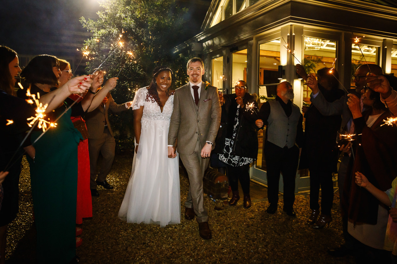 bride and groom walk through tunnel of guests holding sparklers in he evening at relaxed gate_street_barn wedding guildford with candid unposed natural storytelling wedding photography by documentary wedding photographer surrey sussex